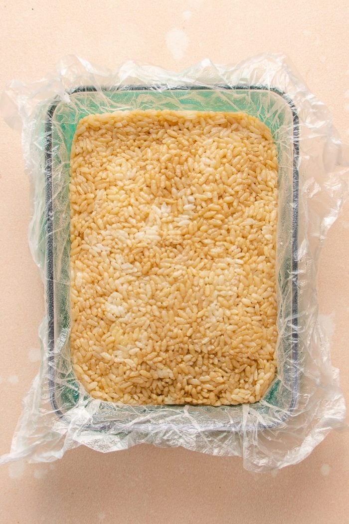 Photo shows seasoned white sushi rice pressed into a block in a glass container lined with saran wrap