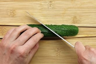 Photo shows two chopsticks on each side of a cucumber with one hand holding the knife at a 45 degree angle cut to slice the cucumber