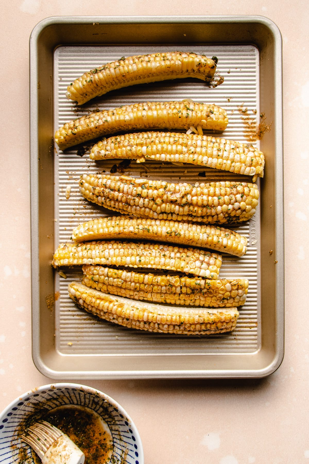 Photo shows corn cobs brushed with butter sauce over a sheet pan