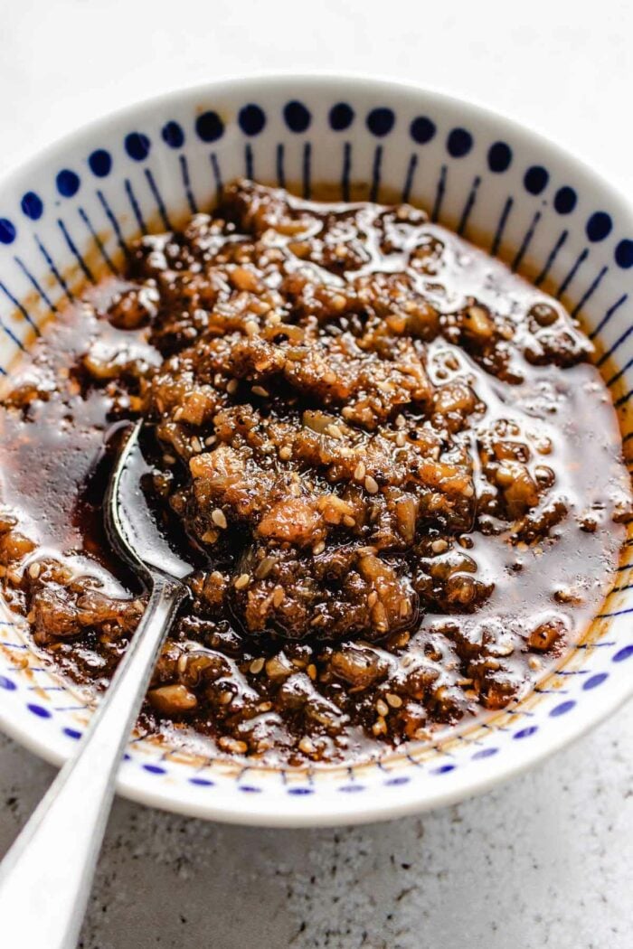 A close side shot photo shows the cooked shacha sauce served in a small white color bowl with spoon on the side