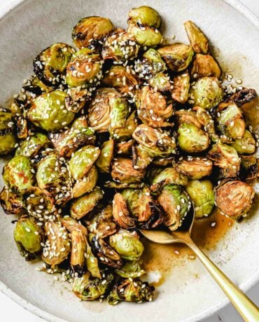 A close shot shows crispy brussels sprouts served on a big white plate with spoon on the side with Asian soy maple glaze