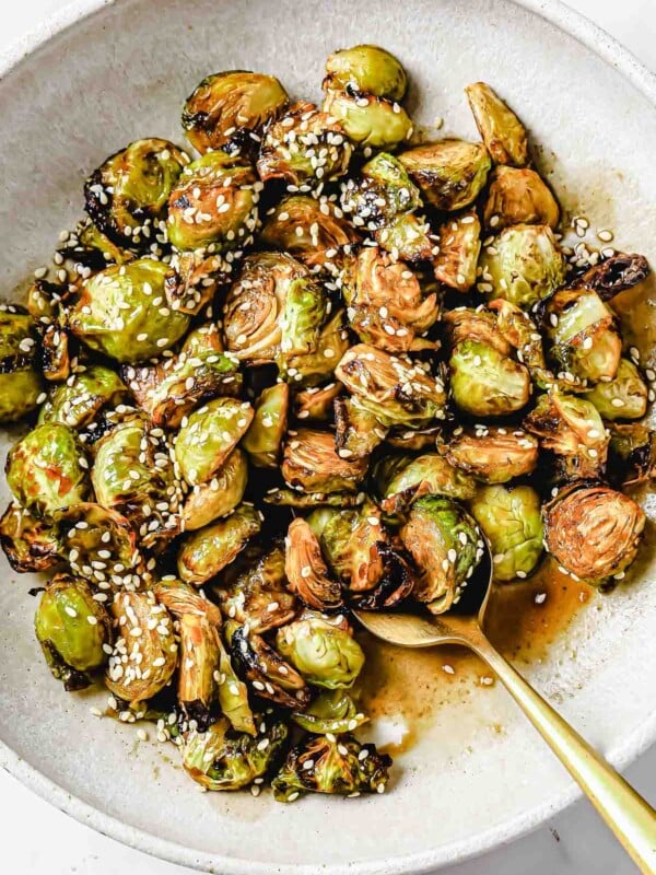 A close shot shows crispy brussels sprouts served on a big white plate with spoon on the side with Asian soy maple glaze