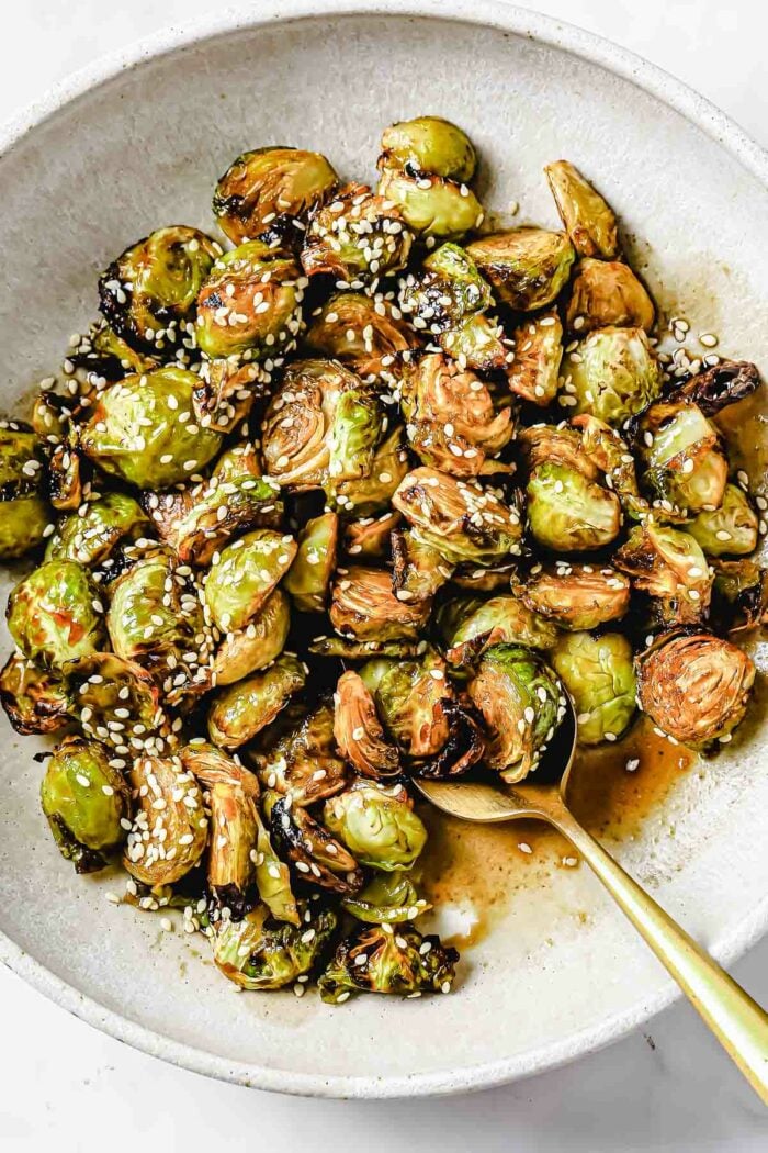 A close shot shows crispy brussels sprouts served on a big white plate with spoon on the side with Asian soy maple glaze