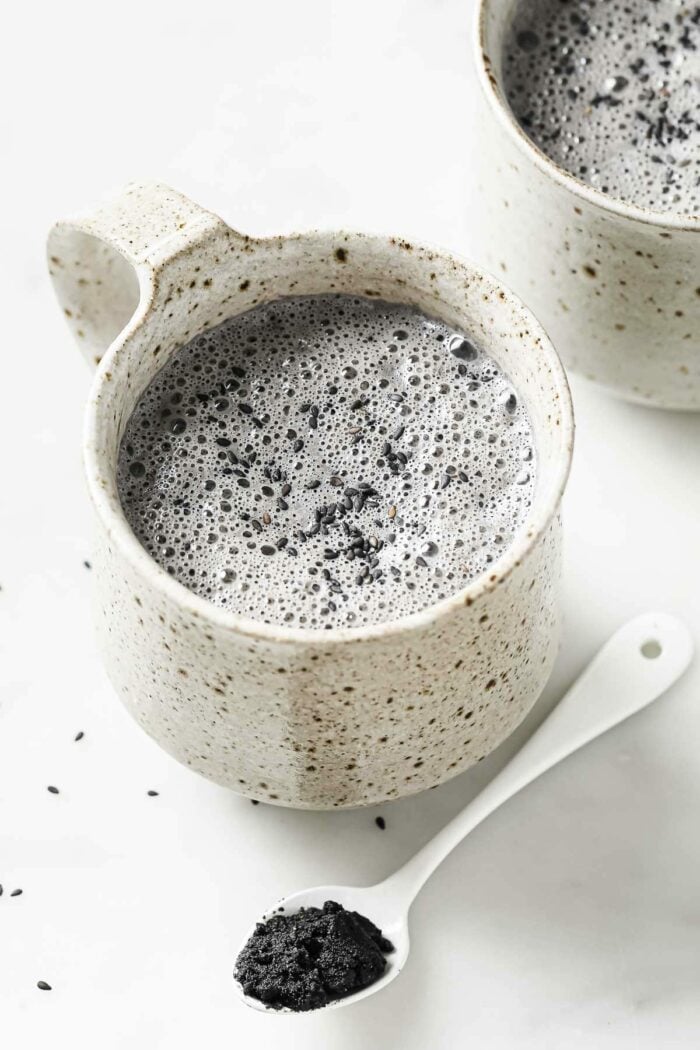 2 white color mugs with black sesame latte milk drink and a small teaspoon of black sesame paste on the side