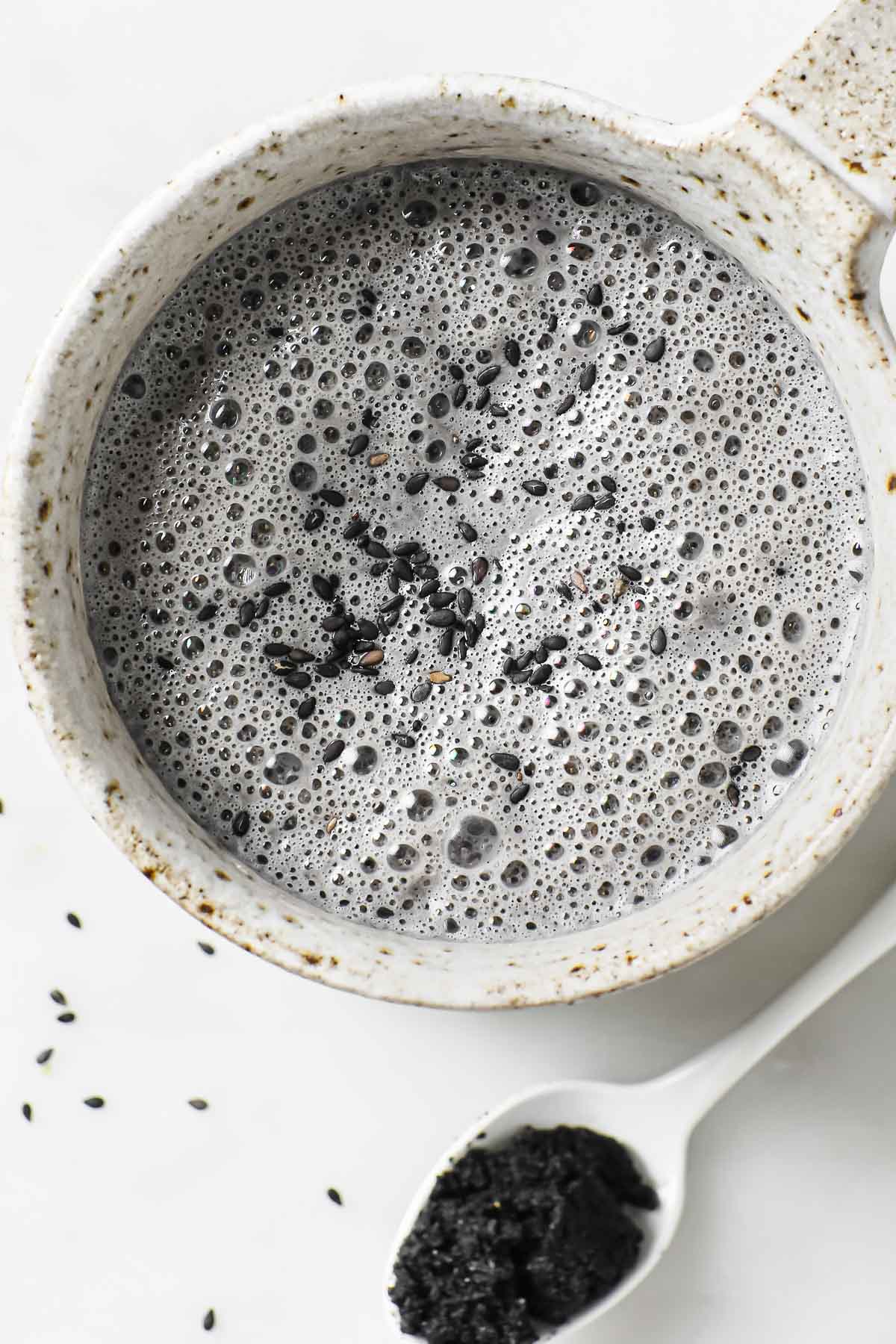Photo shows a white color mug with black sesame milk drink with a teaspoon of sesame paste on the side