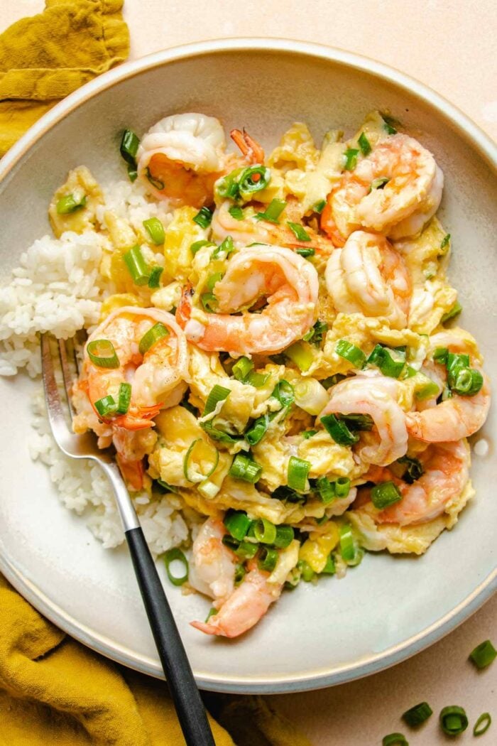 A close shot shows silky scrambled eggs with shrimp served over a bed of steamed white rice on a white plate