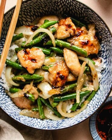 A close shot shows seared chicken breasts sauteed with green beans and onion served with white rice in a blue color bowl