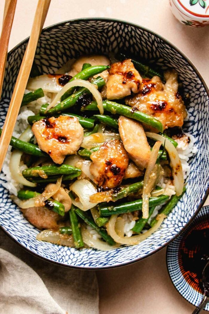 A close shot shows seared chicken breasts sauteed with green beans and onion served with white rice in a blue color bowl