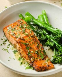 A close shot shows air fried salmon fillet with dijon mustard lemon sauce and broccolini on the side in a white plate