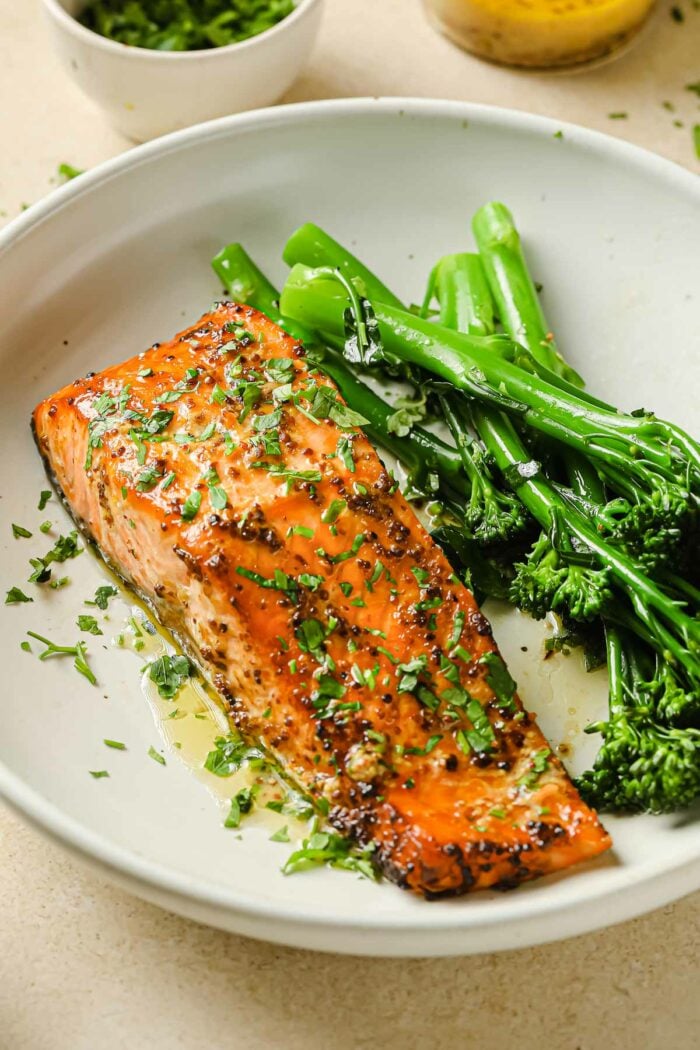 A close shot shows air fried salmon fillet with dijon mustard lemon sauce and broccolini on the side in a white plate