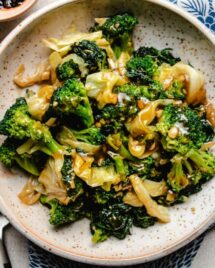 A close shot shows sauteed kale, broccoli, and cabbage served in a neutral color plate with blue napkins on the side.