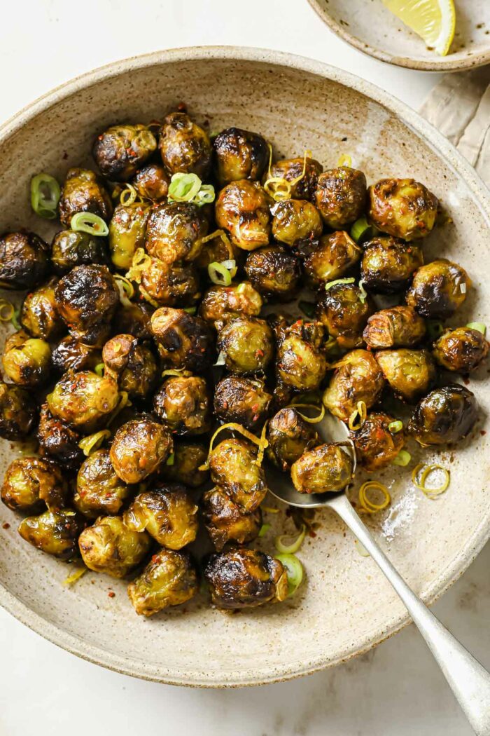An overhead close shot shows brussel sprouts air fried to golden with a little crispy outside served in a neutral color bowl with lemon wedges on the side