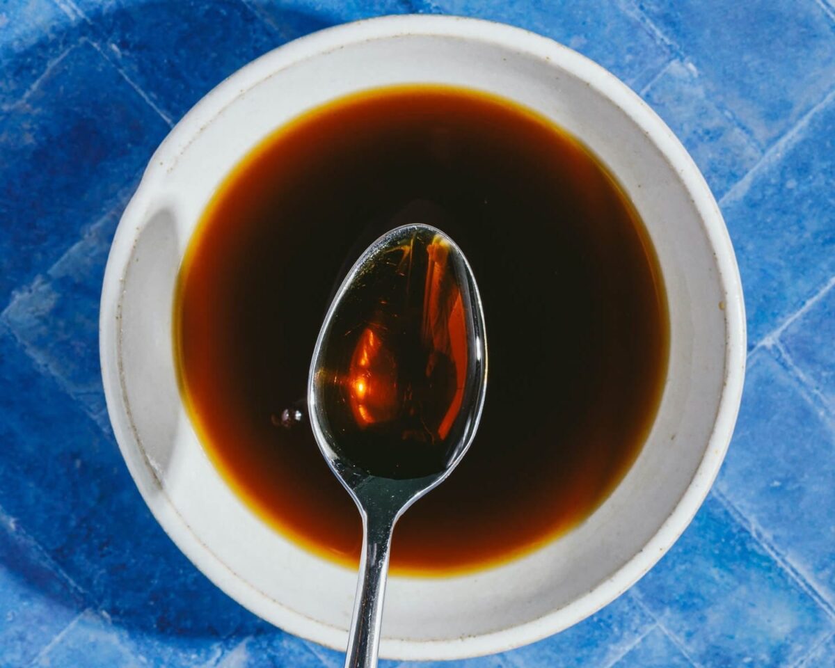 A metal spoon rests in a white bowl filled with homemade teriyaki sauce (gluten-free), set on a blue tile surface—perfect for marinating teriyaki chicken in an air fryer.