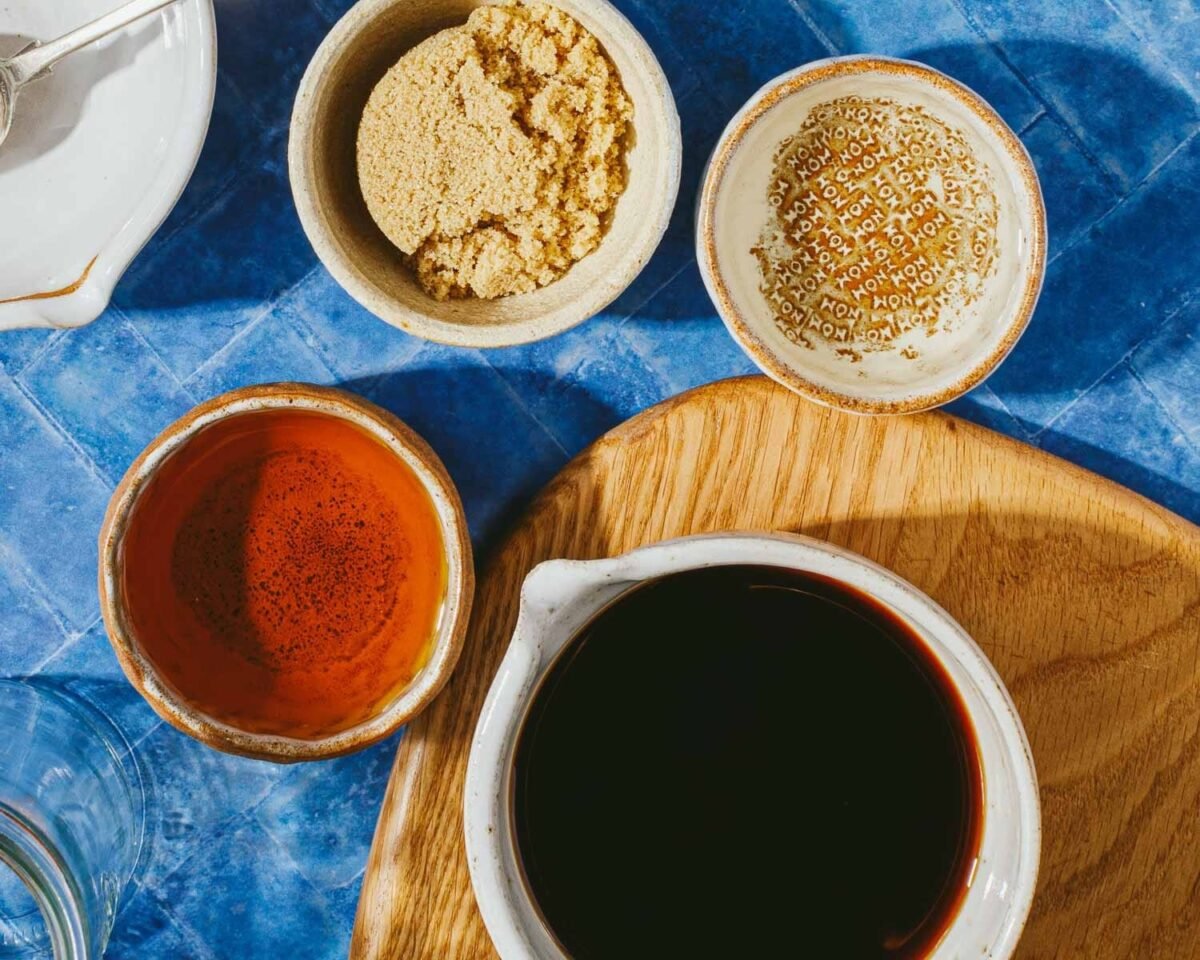 Four containers on a blue and wooden surface, holding brown sugar, mirin, sake, and soy sauce —perfect ingredients for making teriyaki sauce.
