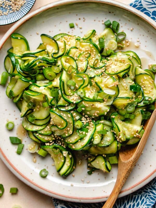 An overhead photo shows thinly sliced and seasoned korean zucchini slices served in a neutral color plate with sesame seeds on top