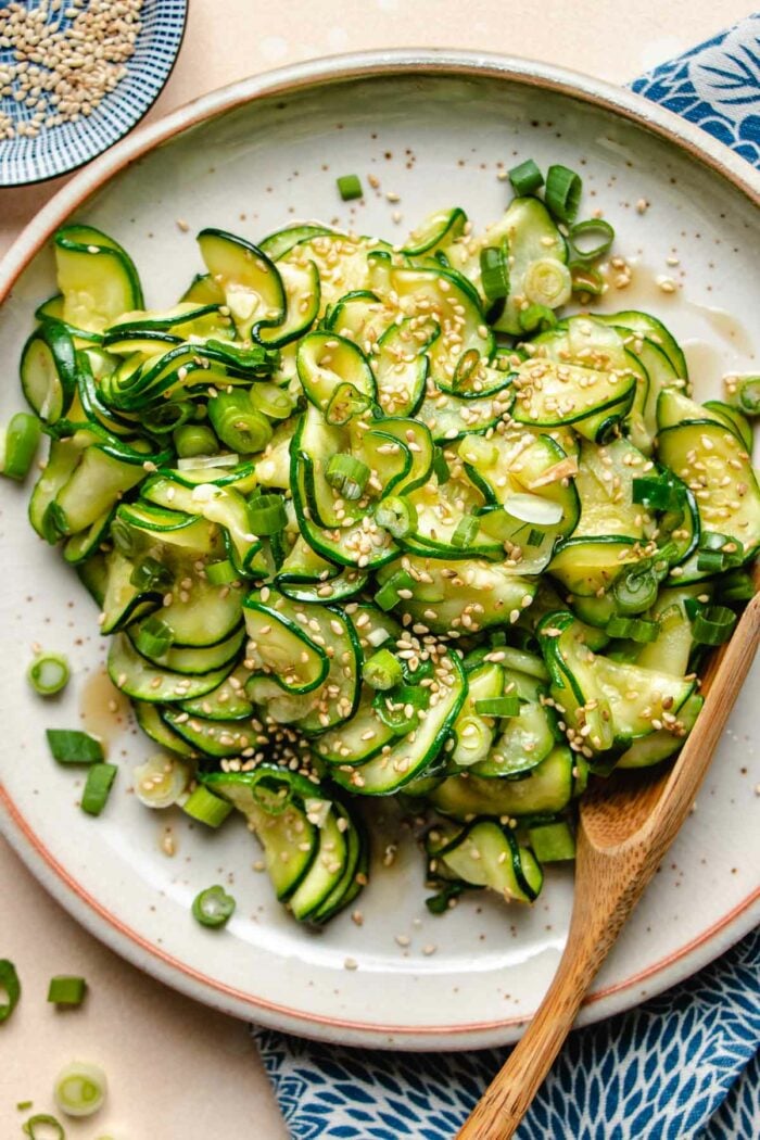 An overhead photo shows thinly sliced and seasoned korean zucchini slices served in a neutral color plate with sesame seeds on top