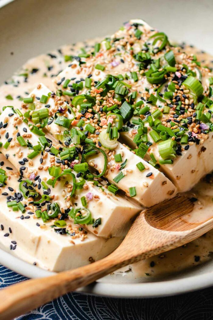 A side close shot shows soft tofu sliced and drizzled with dressing in a plate with a wooden spoon on the side