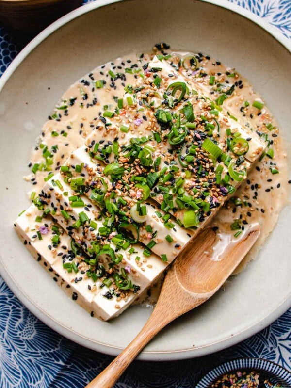 An overhead photo shows silken soft tofu sliced and served in a light gray color plate with dressing and garnish over a blue color napkin