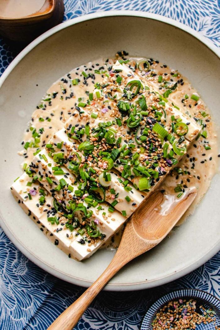 An overhead photo shows silken soft tofu sliced and served in a light gray color plate with dressing and garnish over a blue color napkin