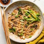 A close shot shows the finishing dish of the peanut noodles, served with extra chili garlic sauce on the side in a white color plate.