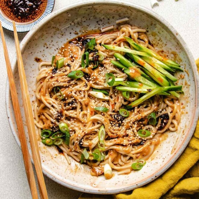 A close shot shows the finishing dish of the peanut noodles, served with extra chili garlic sauce on the side in a white color plate.