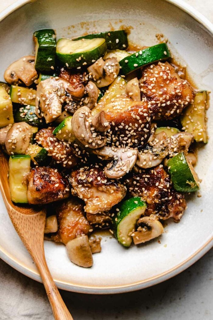 An overhead shot shows crispy chicken chunks with zucchini and mushrooms stir fry served in a white color plate with a wooden spoon.