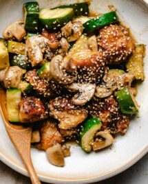 An overhead shot shows crispy chicken chunks with zucchini and mushrooms stir fry served in a white color plate with a wooden spoon.