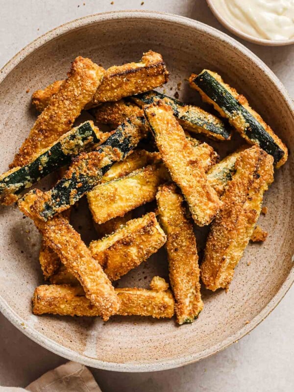 A close shot photo shows crispy zucchini fries served in a neutral color bowl with garlic aioli on the side