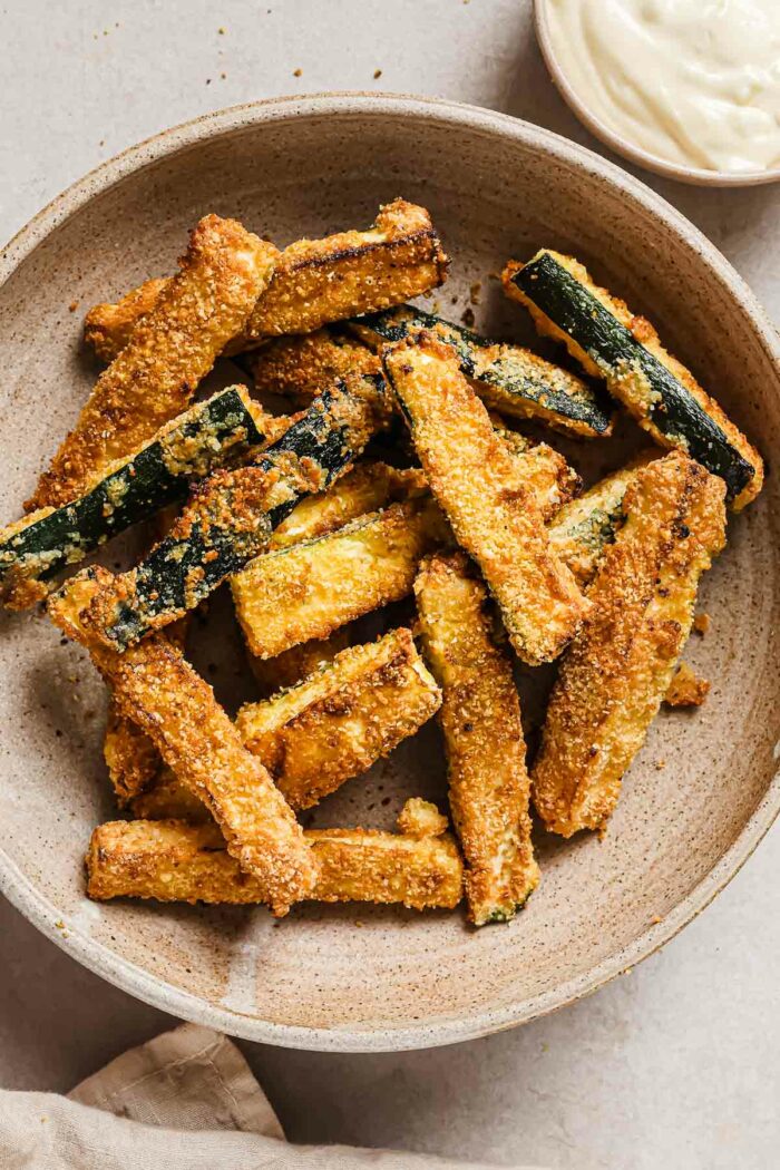 A close shot photo shows crispy zucchini fries served in a neutral color bowl with garlic aioli on the side