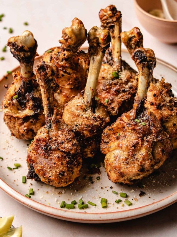 A close shot shows golden crispy chicken lollipop drumsticks served on a neutral color plate with sprinkles on the side.