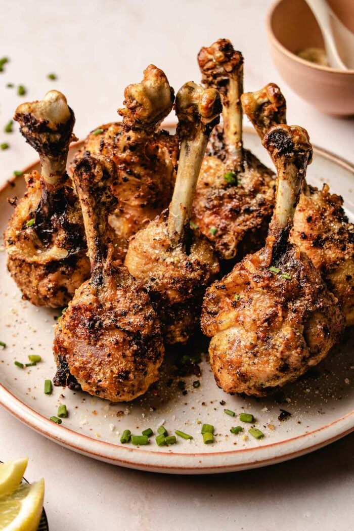 A close shot shows golden crispy chicken lollipop drumsticks served on a neutral color plate with sprinkles on the side.