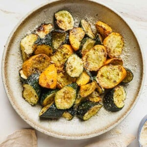 A close shot shows air fryer zucchini squash slices served on a white plate with napkin on the side