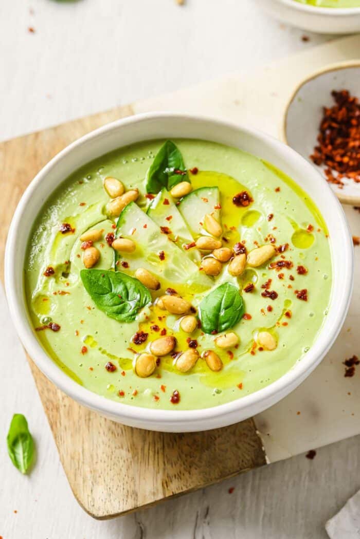 An image shows cucumber gazpacho served in a white bowl with chili pepper flakes on the side.