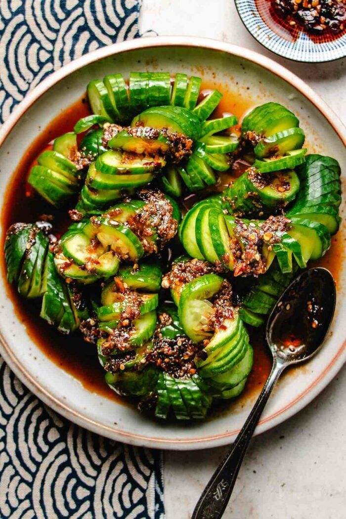 Photo shows the dish Spicy Cucumber Salad spiral-cut served with Chinese chili dressing on a light colored plate with chili sauce on the side.