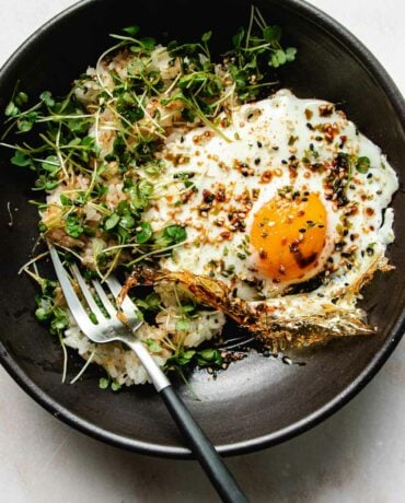 Photo shows an crispy fried egg made in air fryer served on a black plate with rice and garnishes