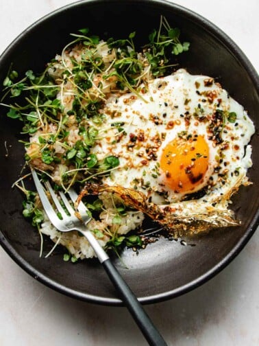 Photo shows an crispy fried egg made in air fryer served on a black plate with rice and garnishes