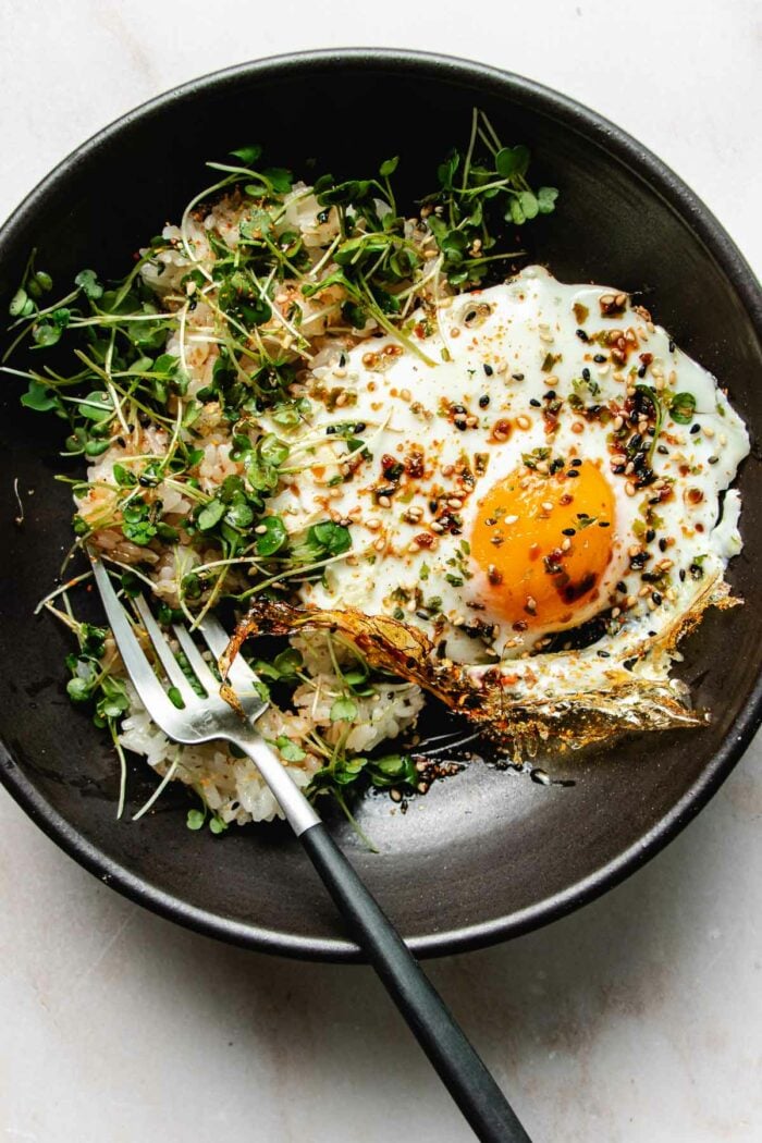 Photo shows an crispy fried egg made in air fryer served on a black plate with rice and garnishes