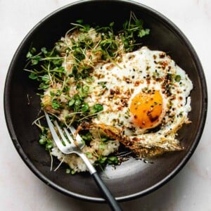 Photo shows a recipe photo for pan fried egg in air fryer with crispy edges served in a black color plate with rice and vegetable garnish