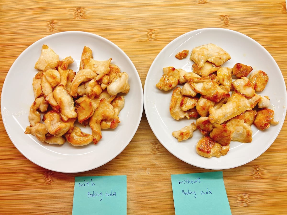Two white plates of cooked chicken breast pieces on a wooden surface; the left plate is labeled with baking soda and the right plate is labeled without baking soda, illustrating how to velvet chicken for a Chinese chicken stir fry marinade by showing the difference in appearance between the two batches.