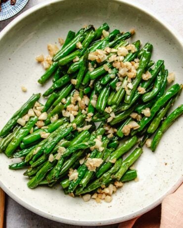 Image shows Din tai fung copycat green beans with crisp exterior and topped with loads of minced garlic served in a white plate.