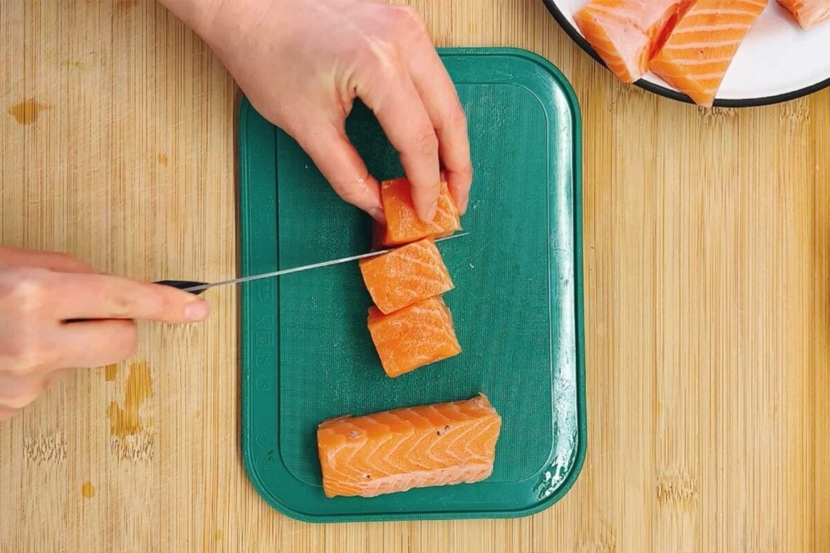 A person slices raw salmon into cubes on a green cutting board, prepping Bang bang salmon bites, with more salmon pieces nearby on a plate, all on a wooden surface.