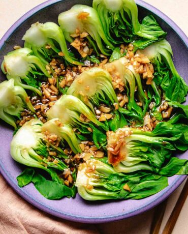 A purple plate filled with steamed baby bok choy with garlic and oyster sauce drizzled on top.