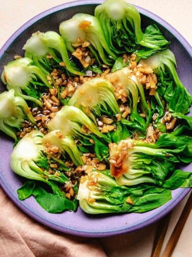 A purple plate filled with steamed baby bok choy with garlic and oyster sauce drizzled on top.