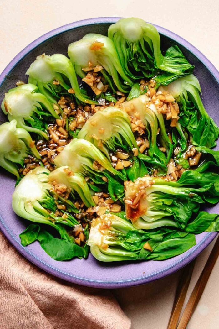 A purple plate filled with steamed baby bok choy with garlic and oyster sauce drizzled on top.