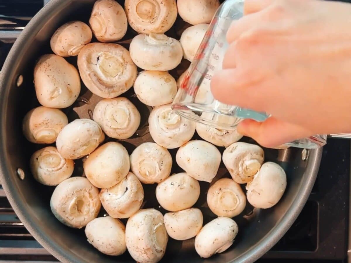 A hand pours liquid from a measuring cup into a pan filled with whole white mushrooms on a stovetop, preparing steak house mushrooms for the perfect savory side.