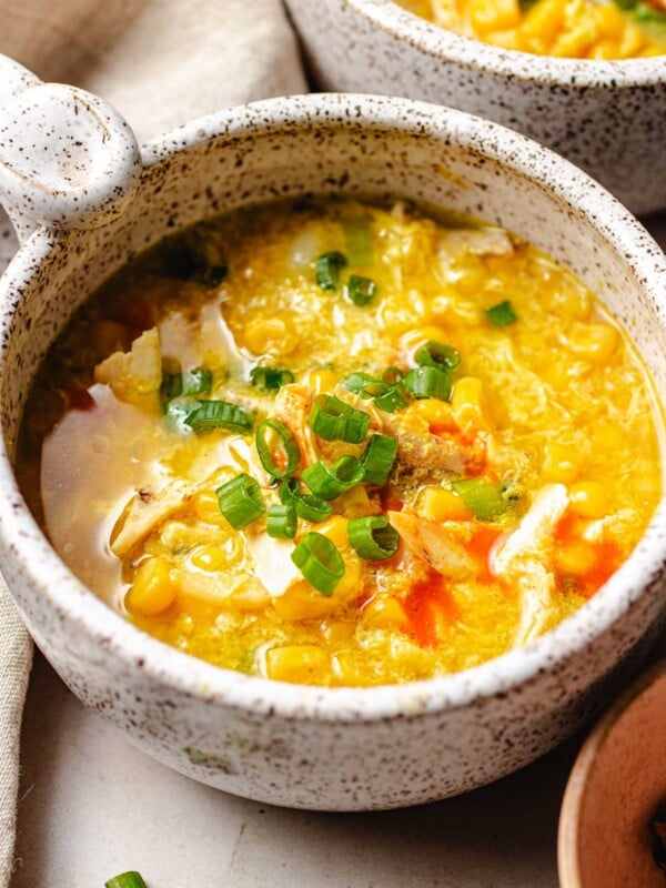 Photo shows chicken corn egg drop soup served in two white soup bowls with spring onion and chili sauce garnish on top.