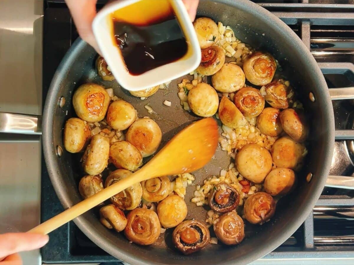 A person pours sauce from a small white container into a skillet with whole mushrooms and chopped onions, stirring with a wooden spoon—a classic step in making steakhouse mushrooms on the stovetop.
