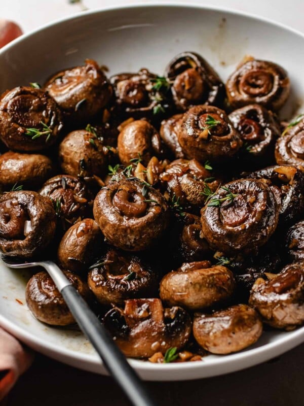 Image shows sauteed steakhouse mushrooms served in a white plate with thyme garnish.