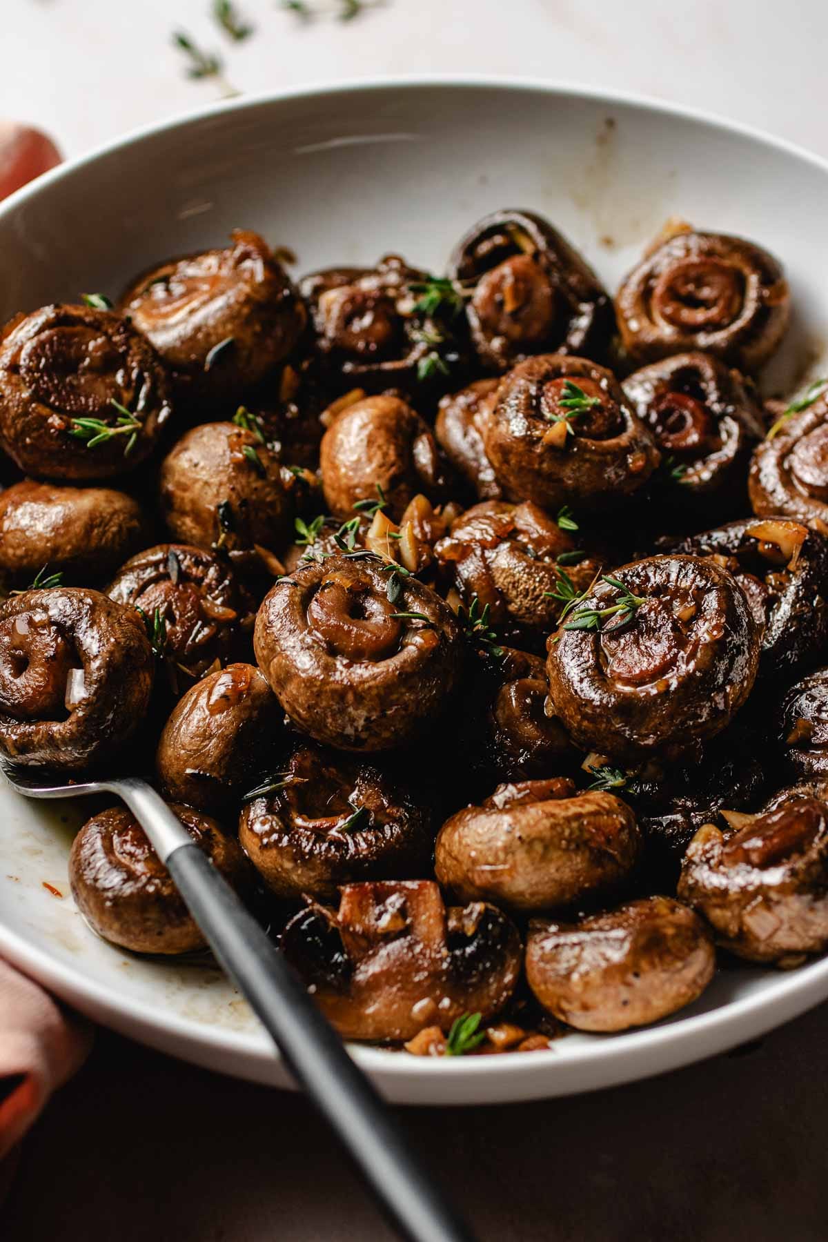 Image shows sauteed steakhouse mushrooms served in a white plate with thyme garnish.