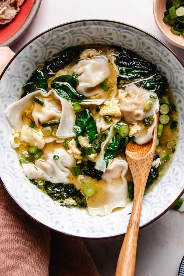 Feature image shows silky soft wontons boiled and served in a bowl of egg drop soup with spinach and seaweed in the bowl.
